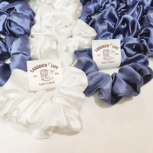 May include: Scrunchies in white and periwinkle blue satin fabric. The scrunchies are arranged in a group, with some featuring a small tag that reads "LASSOED FOR LIFE" with a boot graphic and "SARA'S BACH".