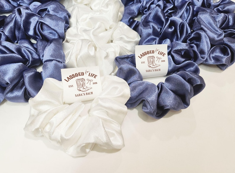 May include: Scrunchies in white and periwinkle blue satin fabric. The scrunchies are arranged in a group, with some featuring a small tag that reads "LASSOED FOR LIFE" with a boot graphic and "SARA'S BACH".
