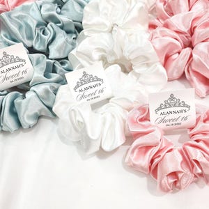 May include: Three satin scrunchies in light blue, white, and pink with a personalized tag that reads "Alannah's Sweet 16 04.19.2023".