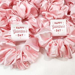 May include: Pink satin scrunchies with white labels that say "Happy Galentine's Day" with pink hearts.