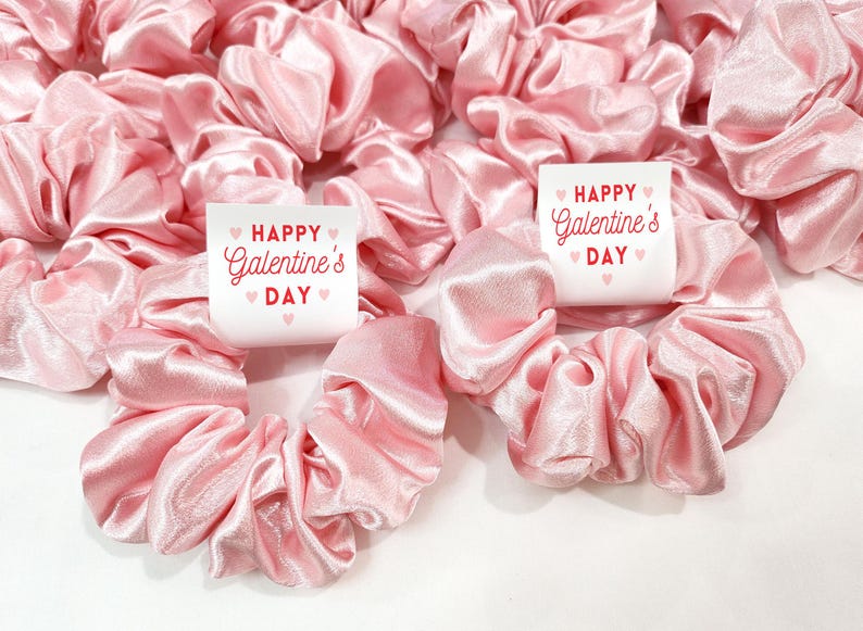 May include: Pink satin scrunchies with white labels that say "Happy Galentine's Day" with pink hearts.