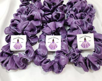 May include: A collection of purple satin scrunchies, each with a small tag. The tags feature a graphic of a girl in a purple dress and the text "Eliana's Quinceañera SEP 14 2025". The scrunchies are arranged on a white surface.