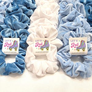 May include: A collection of velvet scrunchies in shades of blue and white. Some scrunchies have a small card that says "Let's Roll" with a roller skate graphic and "Chloe's 8th Birthday". These hair accessories are perfect for a birthday party.