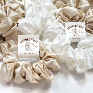 May include: A collection of satin scrunchies in shades of cream and champagne. The scrunchies are arranged in a pile, with a tag that reads "Lassoed for Life" and "Rox's Bach" with a boot and bow graphic.