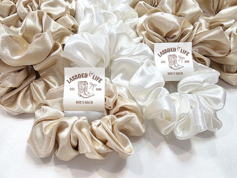 May include: A collection of satin scrunchies in shades of cream and champagne. The scrunchies are arranged in a pile, with a tag that reads "Lassoed for Life" and "Rox's Bach" with a boot and bow graphic.