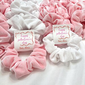 May include: A collection of pink and white velvet scrunchies. Some scrunchies have a small card that reads "happy galentine's day" with the words "love, olivia". These hair accessories are perfect for Valentine's Day.