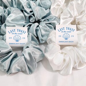 May include: Two sets of hair scrunchies, one set in light blue satin and one set in white satin. Each set has a small card attached with the text "LAST TOAST on the coast 20 25 EMMALEE'S BACH".