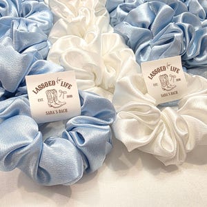 May include: A collection of light blue and white satin scrunchies. Each scrunchie is adorned with a tag that reads "LASSOED for LIFE" with a boot and bow graphic. The tags also include "EST. 2018" and "SARA'S RANCH".
