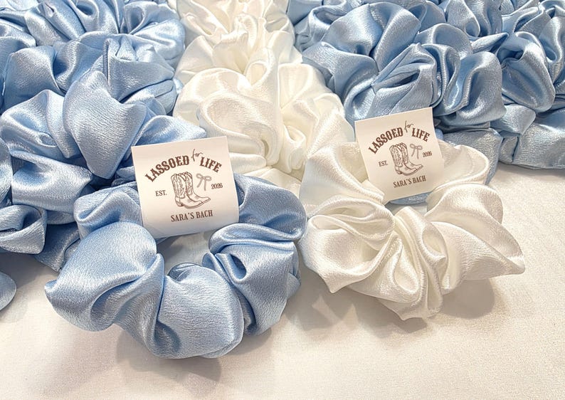 May include: A collection of light blue and white satin scrunchies. Each scrunchie is adorned with a tag that reads "LASSOED for LIFE" with a boot and bow graphic. The tags also include "EST. 2018" and "SARA'S RANCH".