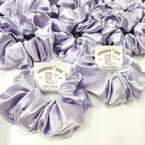 May include: A collection of light purple satin scrunchies. The scrunchies are gathered and ruffled, with a shiny, smooth texture. Each scrunchie is next to a small white tag with the text "LASSOED LIFE EST. 2020 SARA'S BACH" and a drawing of cowboy boots.