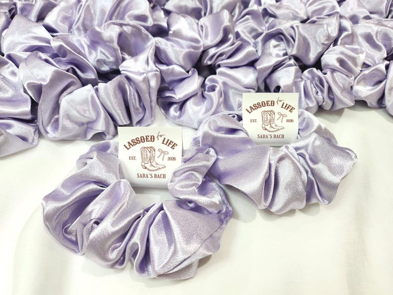 May include: A collection of light purple satin scrunchies. The scrunchies are gathered and ruffled, with a shiny, smooth texture. Each scrunchie is next to a small white tag with the text "LASSOED LIFE EST. 2020 SARA'S BACH" and a drawing of cowboy boots.
