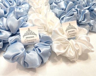 May include: A collection of satin scrunchies in light blue and white. Each scrunchie has a tag with a silver crown design and the text "ANALISA'S Quinceañera 5•16•2026". The scrunchies are bunched together, showcasing their soft, gathered fabric.