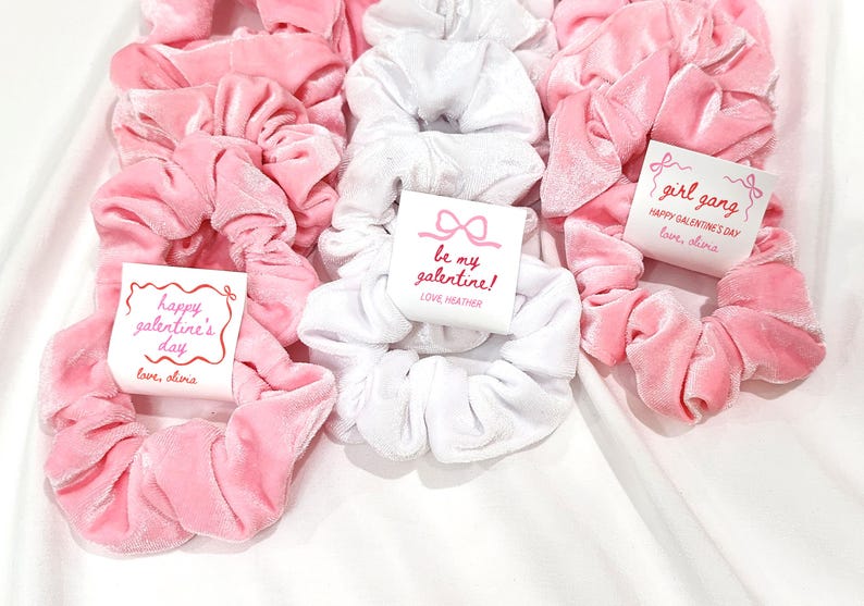 May include: A collection of pink and white velvet scrunchies, some with attached tags. The tags feature text like "Happy Galentine's Day" and "be my galentine!" in pink script. The scrunchies are arranged on a white surface.
