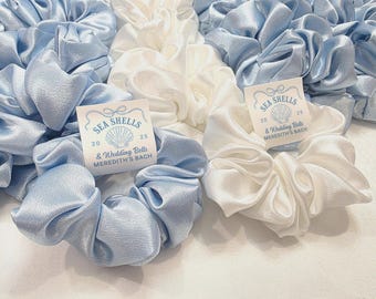 May include: A collection of light blue and white satin scrunchies. Each scrunchie is adorned with a tag that reads "SEA SHELLS & Wedding Bells MEREDITH'S BACH". The scrunchies are bunched together, showcasing their soft, gathered texture.