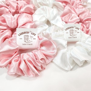 May include: A collection of pink and white satin scrunchies. Some scrunchies have a label that reads "LASSOED for LIFE" with a boot and bow graphic, and "SARA'S BACH" text. The scrunchies are bunched together, creating a soft, textured appearance.