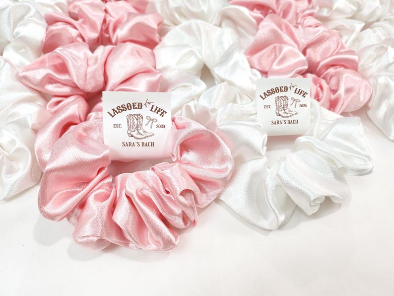 May include: A collection of pink and white satin scrunchies. Some scrunchies have a label that reads "LASSOED for LIFE" with a boot and bow graphic, and "SARA'S BACH" text. The scrunchies are bunched together, creating a soft, textured appearance.