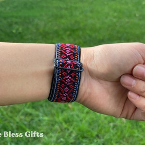 May include: A black elastic watch band featuring a woven pattern in vibrant shades of red, pink, orange, and blue. The band includes a black metal sliding buckle for adjustment, showcasing a detailed geometric design across the fabric surface.