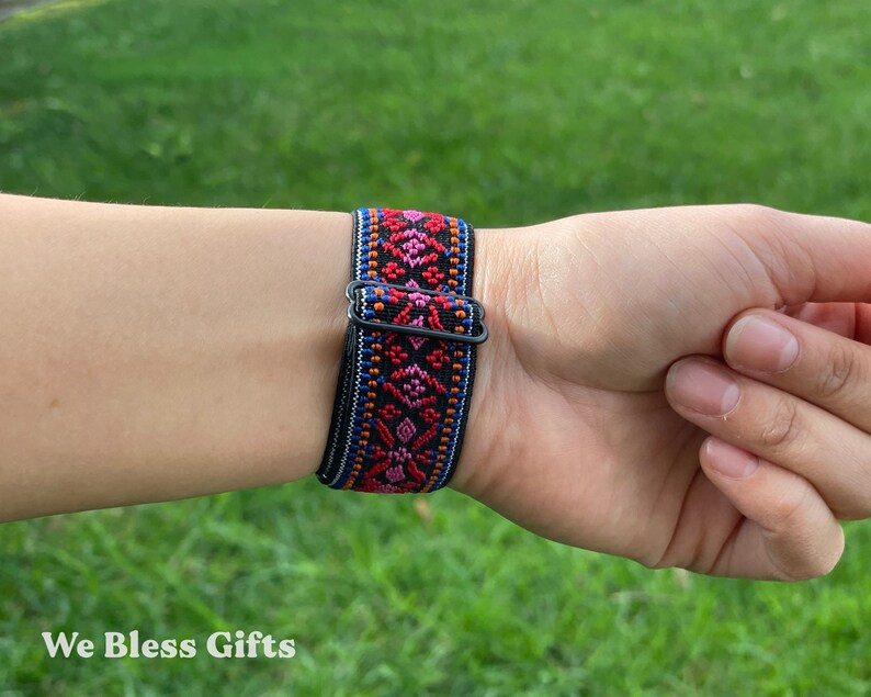 May include: A black elastic watch band featuring a woven pattern in vibrant shades of red, pink, orange, and blue. The band includes a black metal sliding buckle for adjustment, showcasing a detailed geometric design across the fabric surface.
