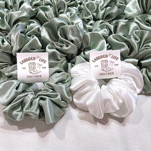 May include: A collection of sage green and white satin scrunchies. One white scrunchie is in the foreground with a label that reads "LASSOED for LIFE" and "SARA'S BACH". The scrunchies are gathered and ruffled, creating a voluminous look.