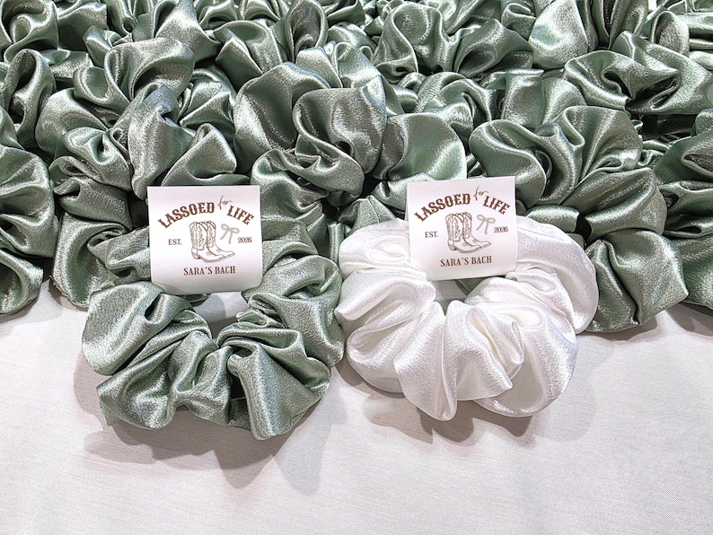 May include: A collection of sage green and white satin scrunchies. One white scrunchie is in the foreground with a label that reads "LASSOED for LIFE" and "SARA'S BACH". The scrunchies are gathered and ruffled, creating a voluminous look.