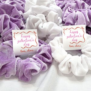May include: A collection of scrunchies in shades of lavender and white. Some scrunchies have a tag that reads "happy galentine's day" and "love, olivia" in red script. The scrunchies are made of a soft, velvety material.