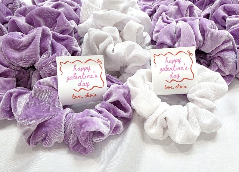 May include: A collection of scrunchies in shades of lavender and white. Some scrunchies have a tag that reads "happy galentine's day" and "love, olivia" in red script. The scrunchies are made of a soft, velvety material.