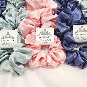 May include: Three satin scrunchies in light blue, pink, and dark blue with personalized tags that read "Alannah's Sweet 16 04.18.2023".