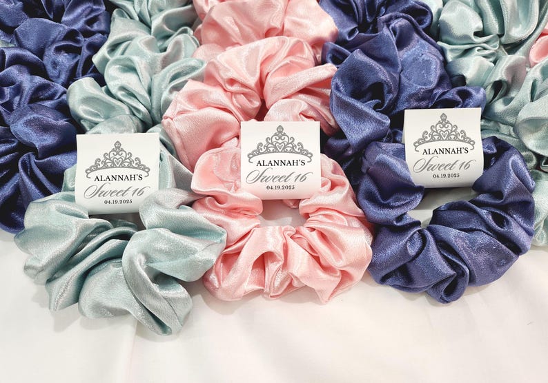 May include: Three satin scrunchies in light blue, pink, and dark blue with personalized tags that read "Alannah's Sweet 16 04.18.2023".