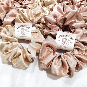May include: A collection of satin scrunchies in shades of champagne, rose, and blush pink. Each scrunchie is gathered and features a tag with the text "LASSOED LIFE EST. 2020 ROX'S RANCH" and a graphic of cowboy boots.