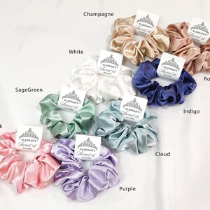 May include: Assortment of ten satin scrunchies in various colors, including pink, sage green, white, champagne, rose gold, indigo, cloud, and purple. Each scrunchie has a small tag attached with the text "Alannah's Sweet 16".