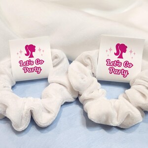 K&ouml;nnte beinhalten: Zwei wei&szlig;e Scrunchies mit rosa und wei&szlig;en Etiketten, auf denen "Let's Go Party" steht, mit einer rosa Silhouette eines Frauenkopfes auf jedem Etikett.