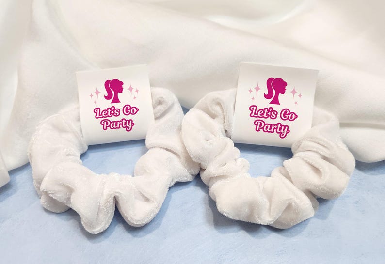 K&ouml;nnte beinhalten: Zwei wei&szlig;e Scrunchies mit rosa und wei&szlig;en Etiketten, auf denen "Let's Go Party" steht, mit einer rosa Silhouette eines Frauenkopfes auf jedem Etikett.