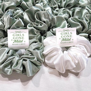 May include: A collection of satin scrunchies in sage green and white. Each scrunchie has a tag that reads "GIRLS GONE Mild" and "REGAN'S BACH" in green text. The scrunchies are grouped together, forming a textured arrangement.