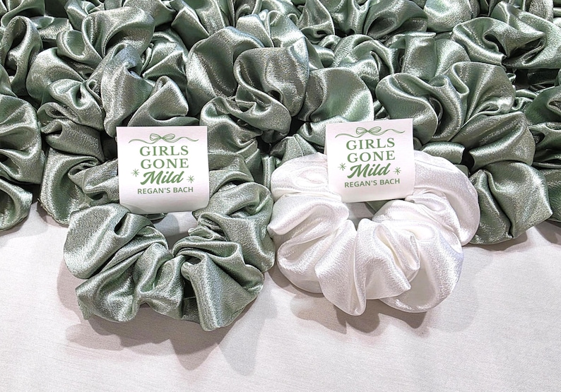 May include: A collection of satin scrunchies in sage green and white. Each scrunchie has a tag that reads "GIRLS GONE Mild" and "REGAN'S BACH" in green text. The scrunchies are grouped together, forming a textured arrangement.