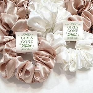May include: A selection of satin scrunchies in blush pink and white. Each scrunchie has a tag that reads "GIRLS GONE Mild" and "Jenn's Bach" in green text. These hair accessories are suitable for a hen party.