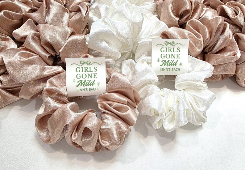May include: A selection of satin scrunchies in blush pink and white. Each scrunchie has a tag that reads "GIRLS GONE Mild" and "Jenn's Bach" in green text. These hair accessories are suitable for a hen party.