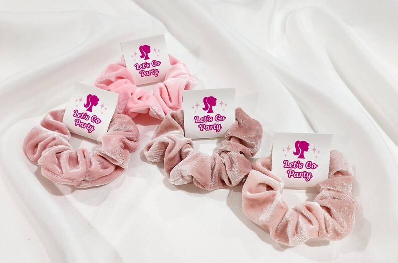 K&ouml;nnte beinhalten: Vier rosafarbene Samt-Haargummis mit wei&szlig;en Etiketten, auf denen "Let's Go Party" mit einer rosa Silhouette eines Frauenkopfes und Sternen steht.