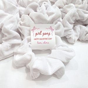 May include: White velvet scrunchies, a popular hair accessory, are scattered across a white surface. A small card with the text "girl gang" and "Happy Galentine's Day" is placed on one of the scrunchies.