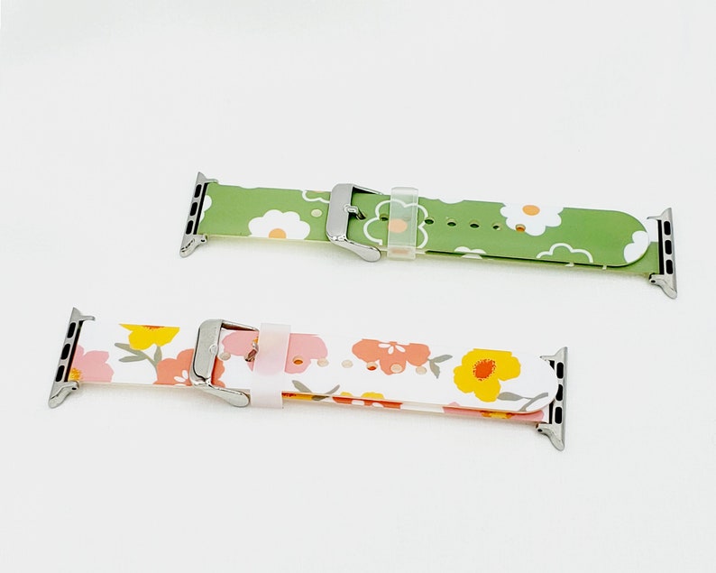 K&ouml;nnte beinhalten: Zwei Apple Watch Armb&auml;nder mit Blumenmustern. Das obere Armband ist gr&uuml;n mit wei&szlig;en Blumen. Das untere Armband ist wei&szlig; mit rosa und gelben Blumen.
