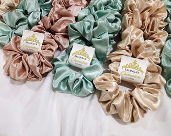 May include: A collection of satin scrunchies in shades of blush, seafoam green, and champagne. Each scrunchie has a small white tag with gold text that reads "Brianna's Quinceañera 12.13.2023".