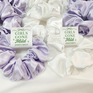 May include: A collection of satin scrunchies in lilac and white. The scrunchies are displayed on a white background. Each scrunchie has a tag with the text "GIRLS GONE Mild" and "Jenn's Bach" in green and black.