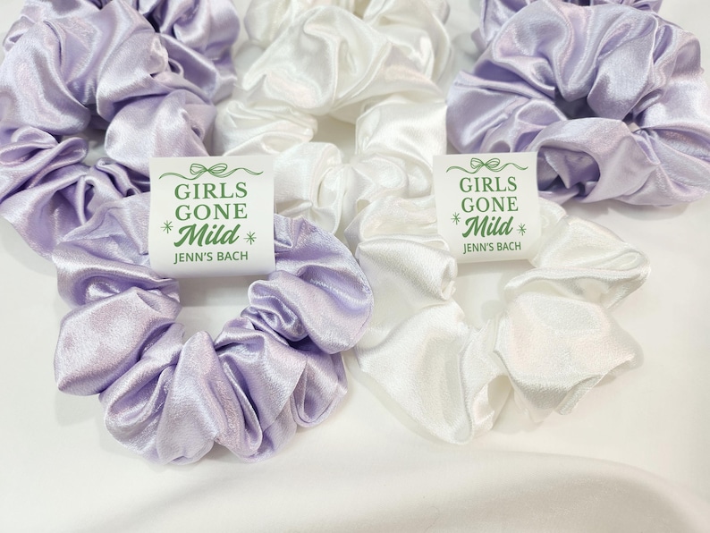 May include: A collection of satin scrunchies in lilac and white. The scrunchies are displayed on a white background. Each scrunchie has a tag with the text "GIRLS GONE Mild" and "Jenn's Bach" in green and black.