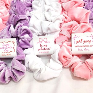 May include: A collection of velvet scrunchies in shades of pink, lavender, and white. Some scrunchies have small cards with text that reads "Happy Galentine's Day", "Be My Galentine!", and "Girl Gang".