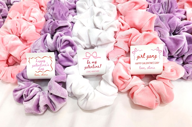 May include: A collection of velvet scrunchies in shades of pink, lavender, and white. Some scrunchies have small cards with text that reads "Happy Galentine's Day", "Be My Galentine!", and "Girl Gang".