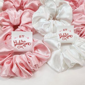 May include: Two satin scrunchies, one pink and one white, with a small card attached to each. The cards have the text "Be my Galentine" in red lettering.