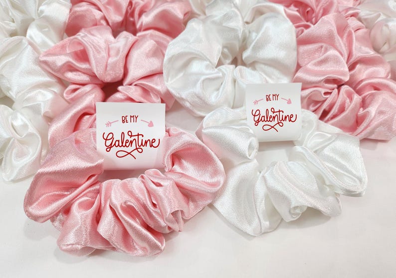 May include: Two satin scrunchies, one pink and one white, with a small card attached to each. The cards have the text "Be my Galentine" in red lettering.