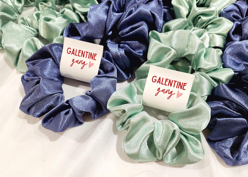 May include: Two sets of satin scrunchies, one set in blue and one set in green, with a white tag that says "Galentine gang" in red lettering with a pink heart.