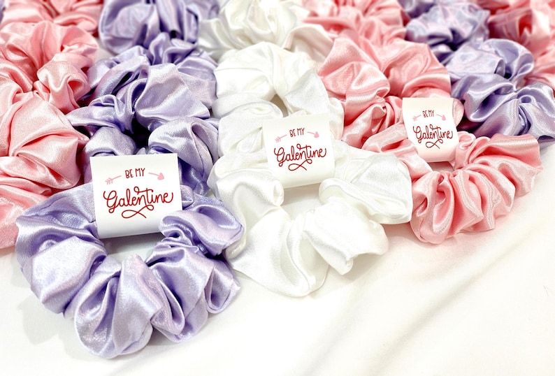 May include: A collection of satin scrunchies in various colors, including lavender, pink, and white. Each scrunchie has a white tag attached with the text "Be My Galentine" in red lettering.