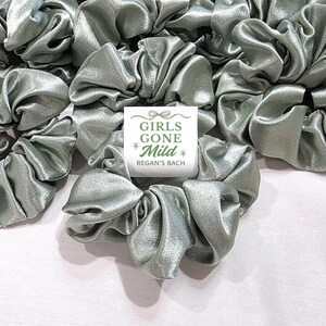 May include: A collection of sage green satin scrunchies, with a shiny, gathered texture, are displayed. A small white card with the text "GIRLS GONE Mild * Regan's Bach" is placed among them. The scrunchies are on a white surface.