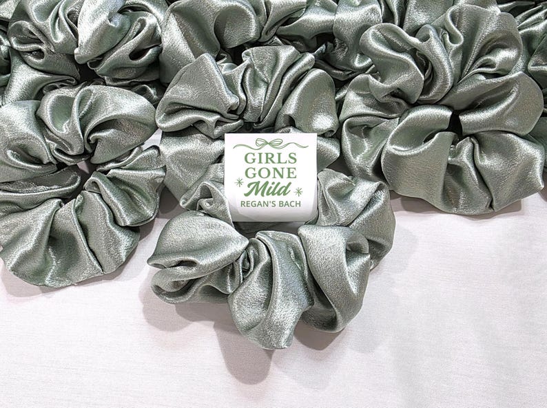 May include: A collection of sage green satin scrunchies, with a shiny, gathered texture, are displayed. A small white card with the text "GIRLS GONE Mild * Regan's Bach" is placed among them. The scrunchies are on a white surface.